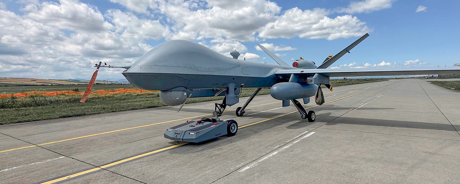 An image of a TowFLEXX TF3 Milspec, a specialized, remote-controlled aircraft tug, in the process of maneuvering an MQ-9 Reaper, a large military drone known for its long-endurance, high-altitude surveillance and reconnaissance capabilities. The TowFLEXX TF3 Milspec is designed for precision, allowing for the safe and efficient handling of aircraft in military applications, demonstrating its utility in facilitating the deployment and maintenance of unmanned aerial systems like the MQ-9 Reaper
